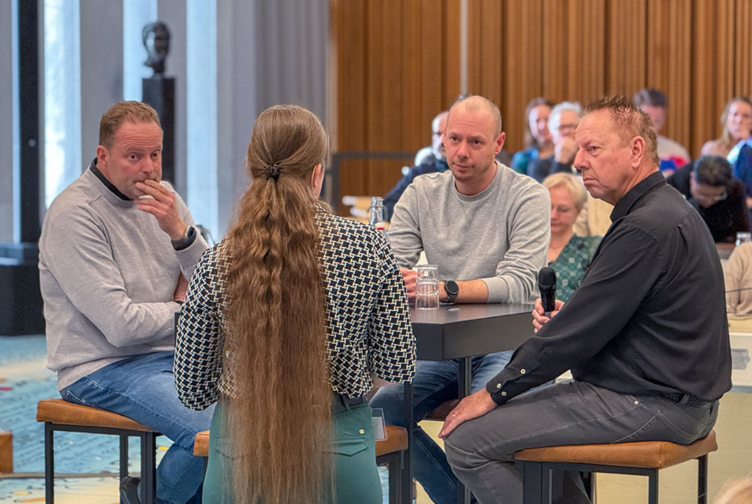 Maurits van de Geijn, Leslie Goedhart en Carel Eltingh zitten van links naar rechts aan een hoge tafel, terwijl ze worden geïnterviewd door Larissa Klaassen die voor hen staat en met de rug naar de camera.