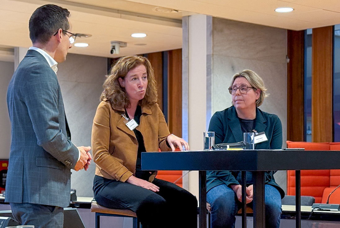 Astrid Oostveen zit aan tafel met zittend rechts naast haar Patricia Bolink. Links staat Michiel Haagen hen te interviewen met de rug naar de cameraks staat